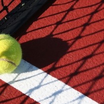 close up of yellow tennis ball on red court in shadow of net